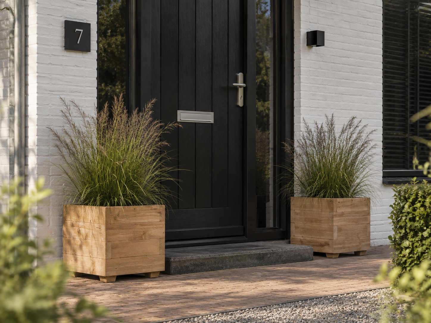 Twee vierkante houten plantenbakken met siergrassen naast een zwarte voordeur van Plant&bak